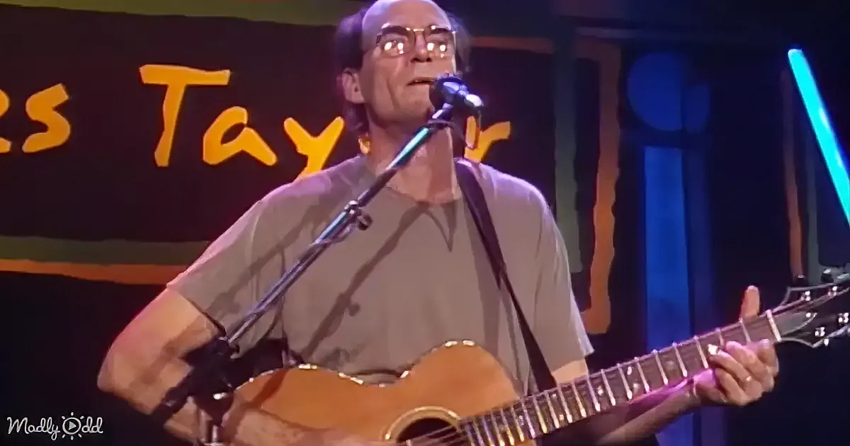 A man in a brown shirt plays an acoustic guitar and sings into a microphone on stage.