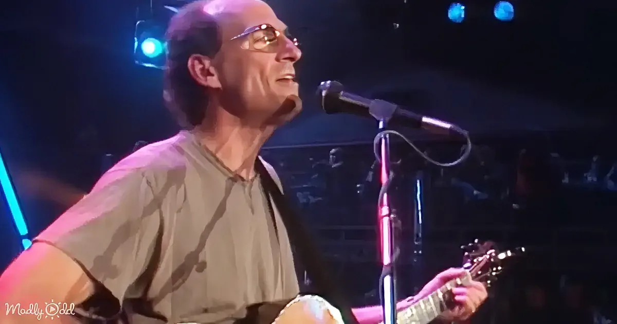 A man in glasses sings into a microphone while playing guitar on stage.