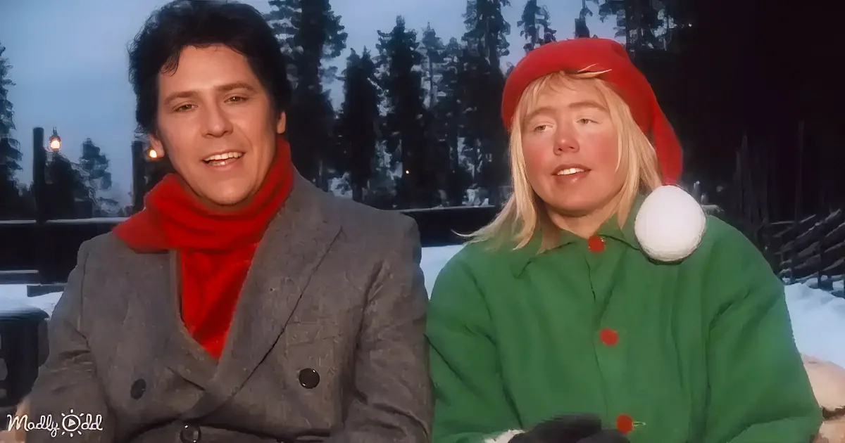 A man in a gray coat and red scarf sits next to a woman in a green elf outfit with a red hat and white pom-pom.