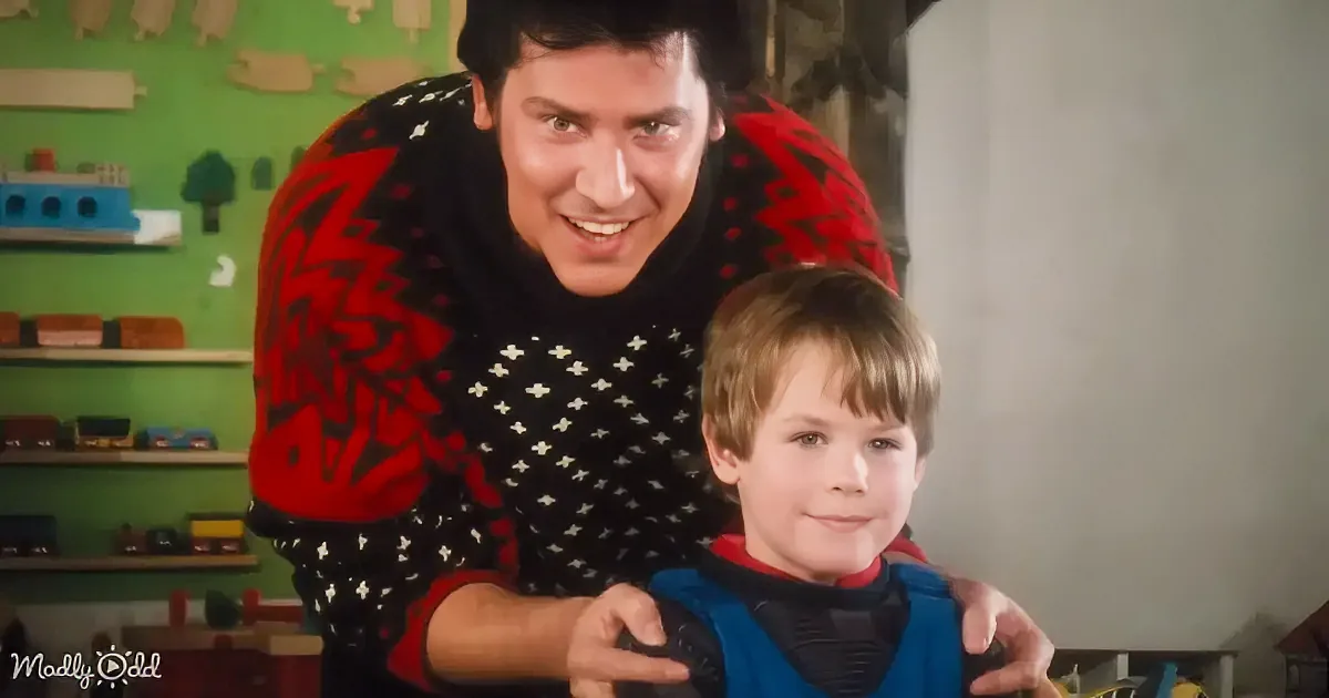 A man in a red and black sweater smiles while holding a young boy in a blue vest, both in a room with shelves of toys.