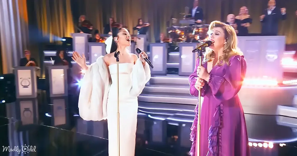 Two women in elegant dresses sing on stage with a live orchestra in the background.