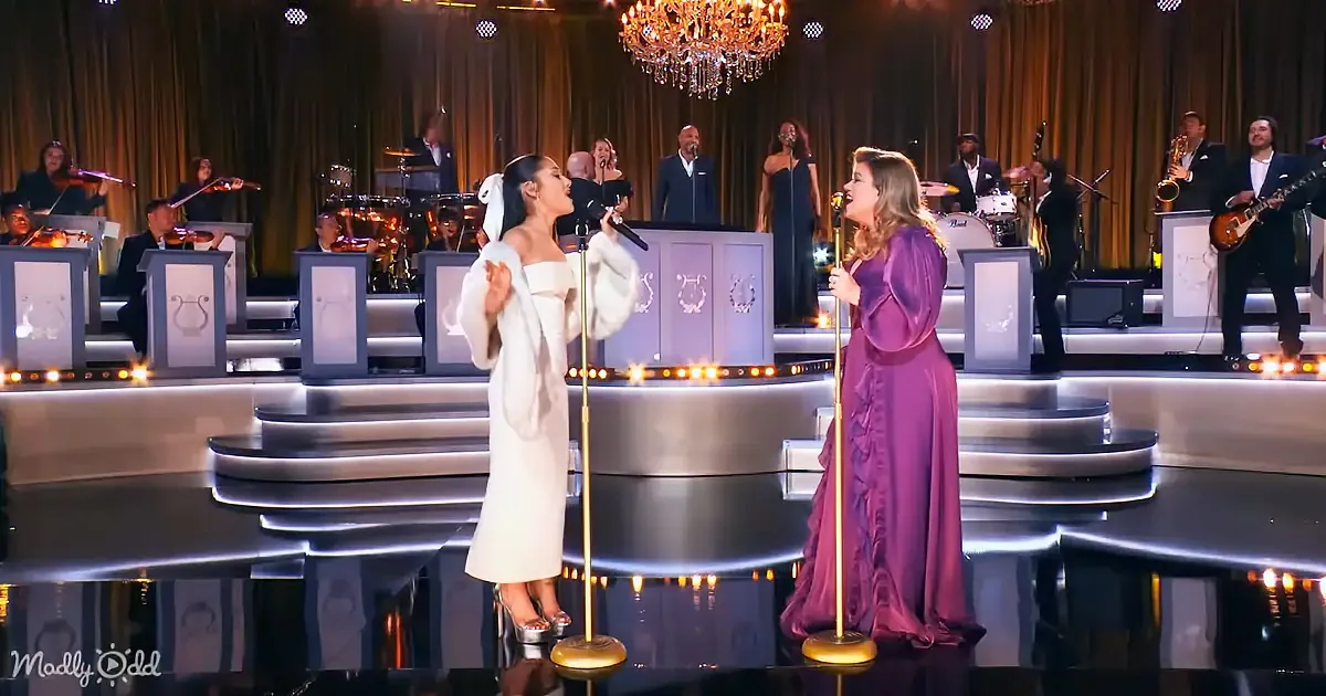 Two women in elegant dresses sing on stage with a band and chandelier backdrop.