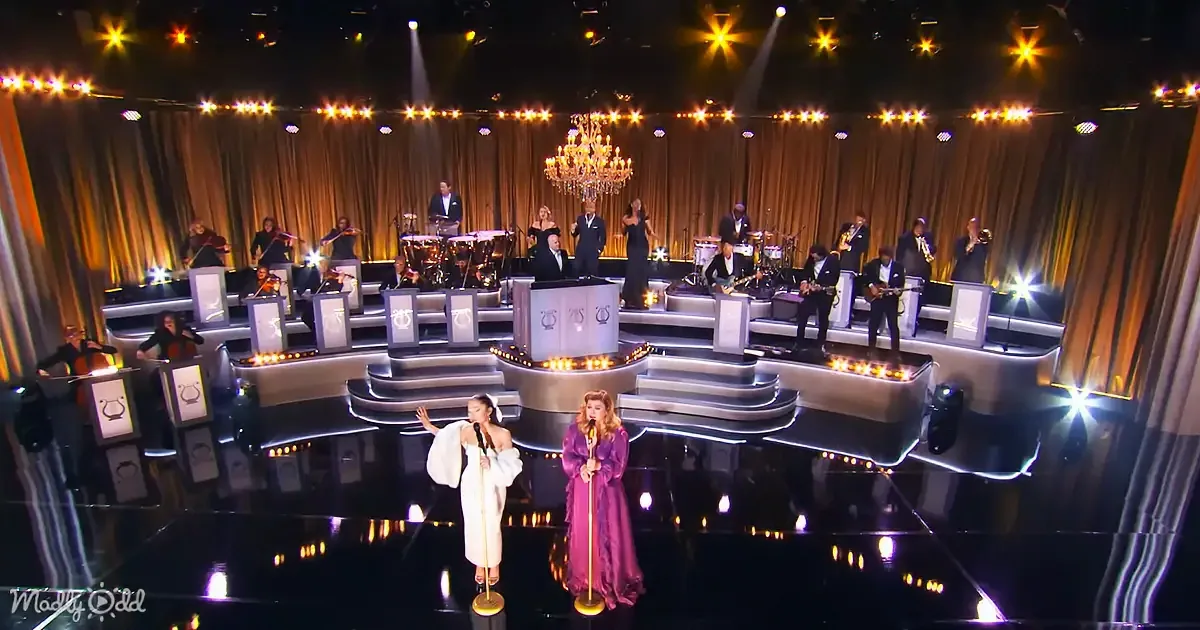Two women in elegant gowns perform on stage with a large orchestra and chandelier backdrop.