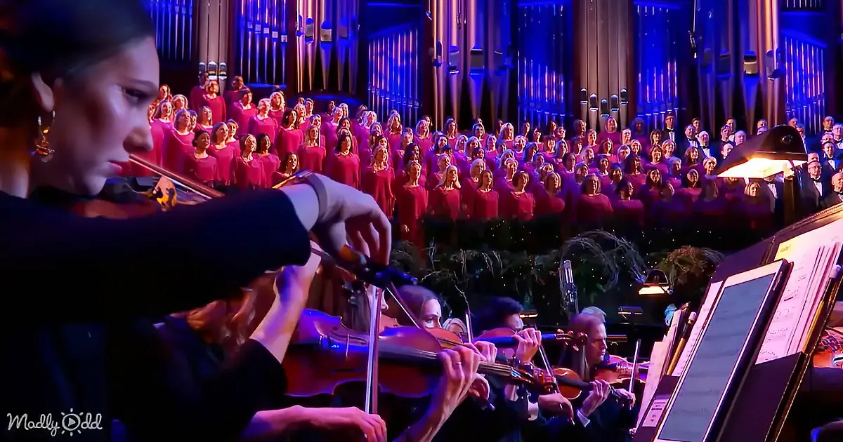 A violinist plays in front of a choir and orchestra in a grand hall.