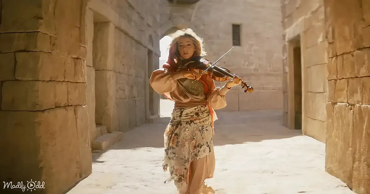 A woman in a floral dress plays a violin in a sunlit stone corridor.