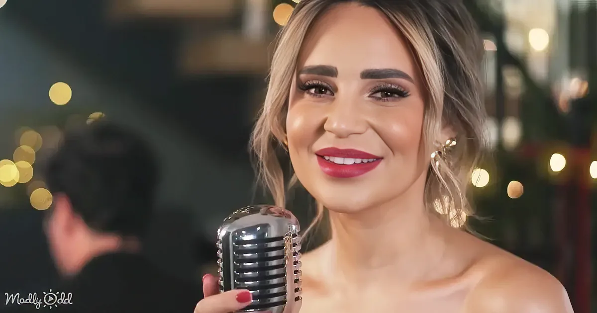 A woman with red lipstick holds a vintage microphone, smiling, with blurred lights and another person in the background.
