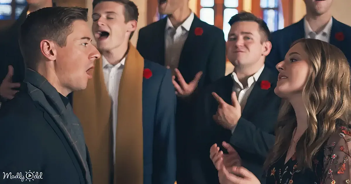 A man in a suit sings passionately, surrounded by a group of people in formal attire, some with red poppies, clapping and...