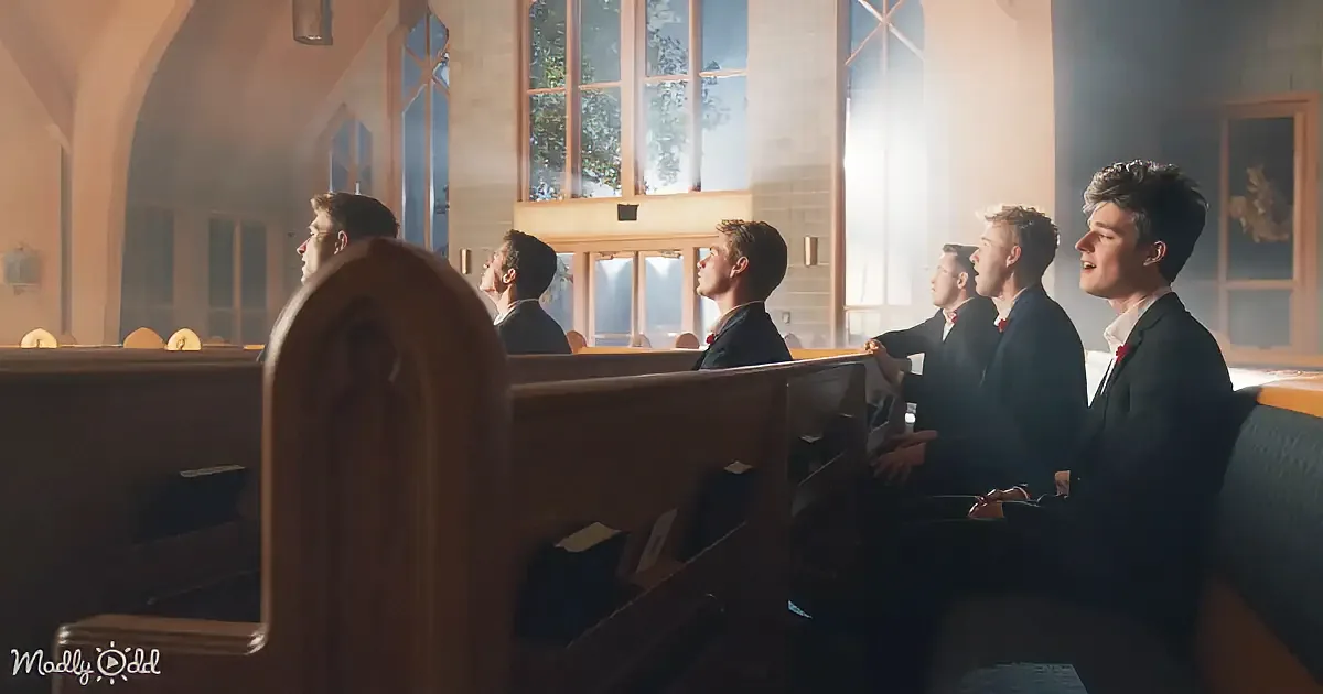Six men in suits sing in a church with sunlight streaming through large windows.