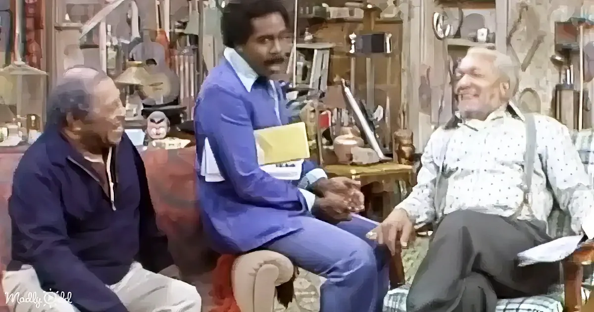 A man in a blue suit sits on a chair, holding a yellow folder, while two others sit beside him in a cluttered room.