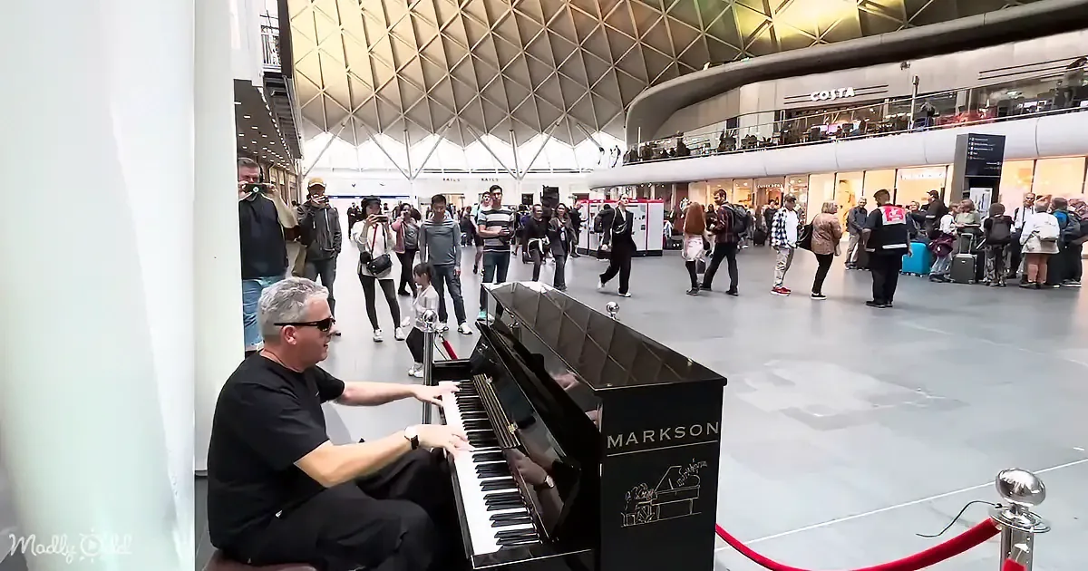 A man plays a Markson piano in a busy train station, surrounded by onlookers.
