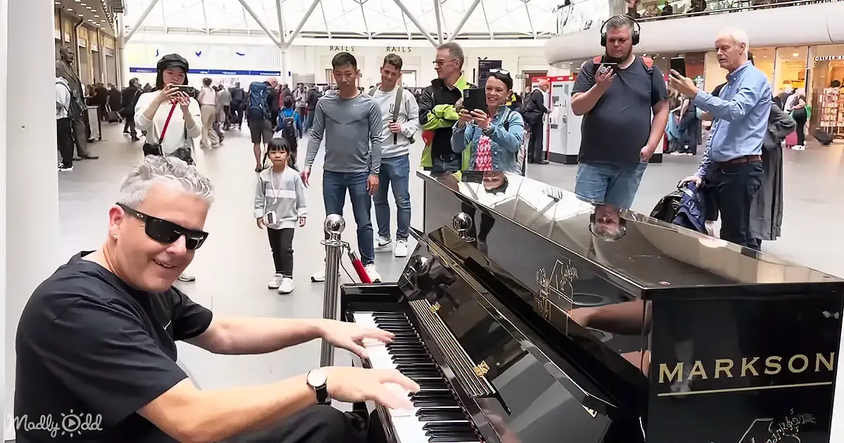 A man in sunglasses plays a Markson piano in a busy airport, surrounded by onlookers taking photos.