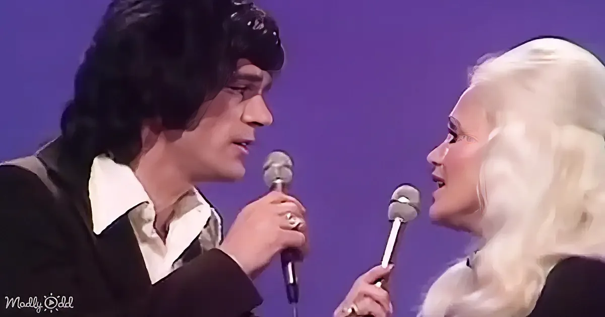 A man and woman sing into microphones, both wearing formal attire, against a purple background.