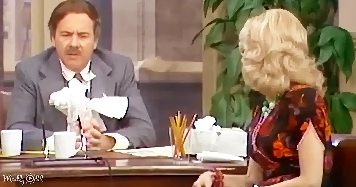 A man in a suit sits at a desk, holding a crumpled piece of paper, while a woman with blonde hair sits across from him.