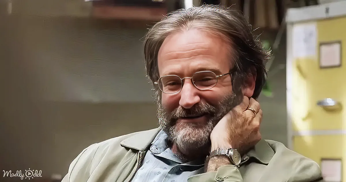 A man with gray hair and beard, wearing glasses and a light jacket, smiles while resting his chin on his hand.