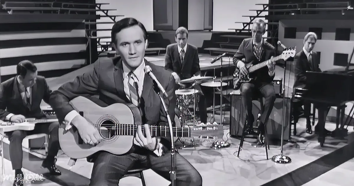 A man in a suit plays guitar on stage with a band, including a drummer and a guitarist.