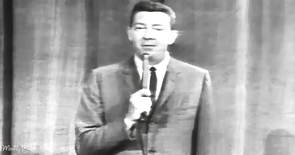 A man in a suit and tie holds a microphone, standing in front of a curtain backdrop.