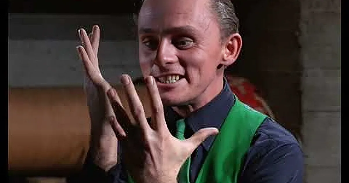 A man in a green suit with question marks, black hat, and a red rose pin stands in a doorway.