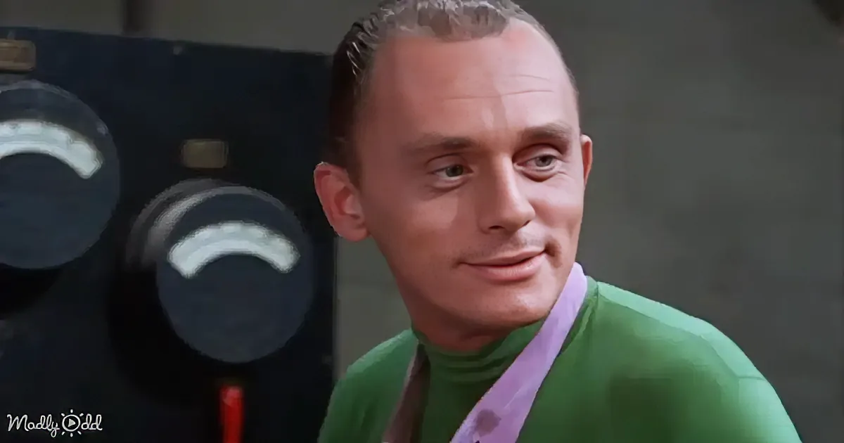 A man in a green turtleneck and purple shirt smiles near a control panel with dials.