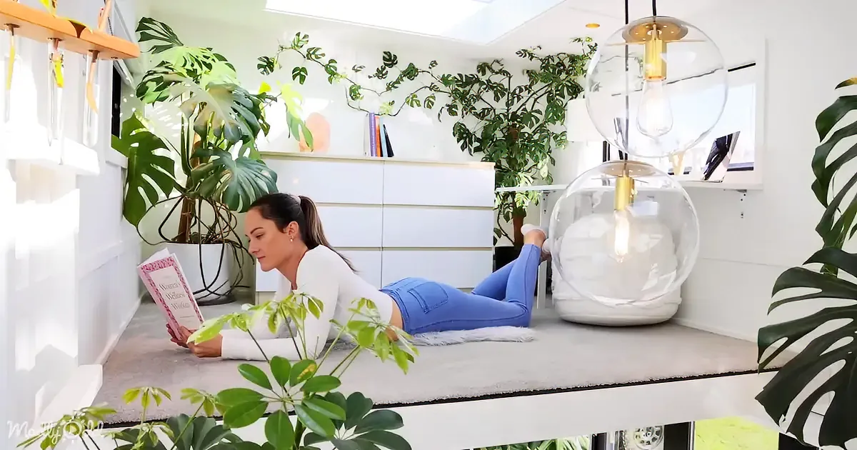 A woman lies on a white rug reading a book in a cozy, plant-filled room with modern lighting.