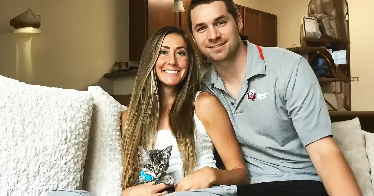 A couple sits on a couch with a kitten, smiling at the camera.