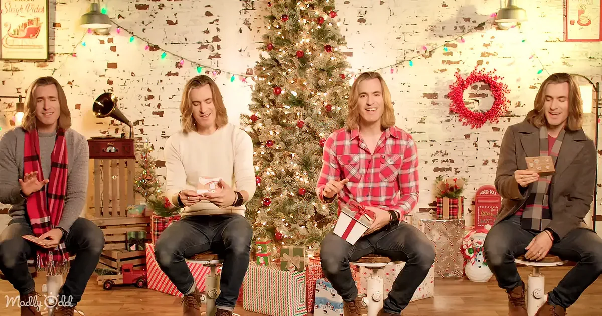 A man with long hair sits in front of a Christmas tree, holding a gift, surrounded by festive decorations.
