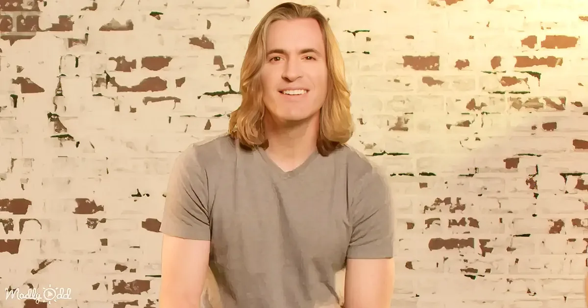 A person with long blonde hair, wearing a gray t-shirt, sits in front of a white brick wall.