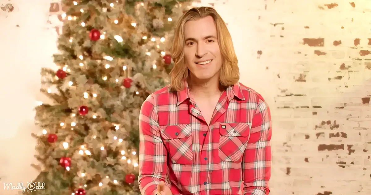 A person in a red plaid shirt smiles in front of a decorated Christmas tree with lights and ornaments.