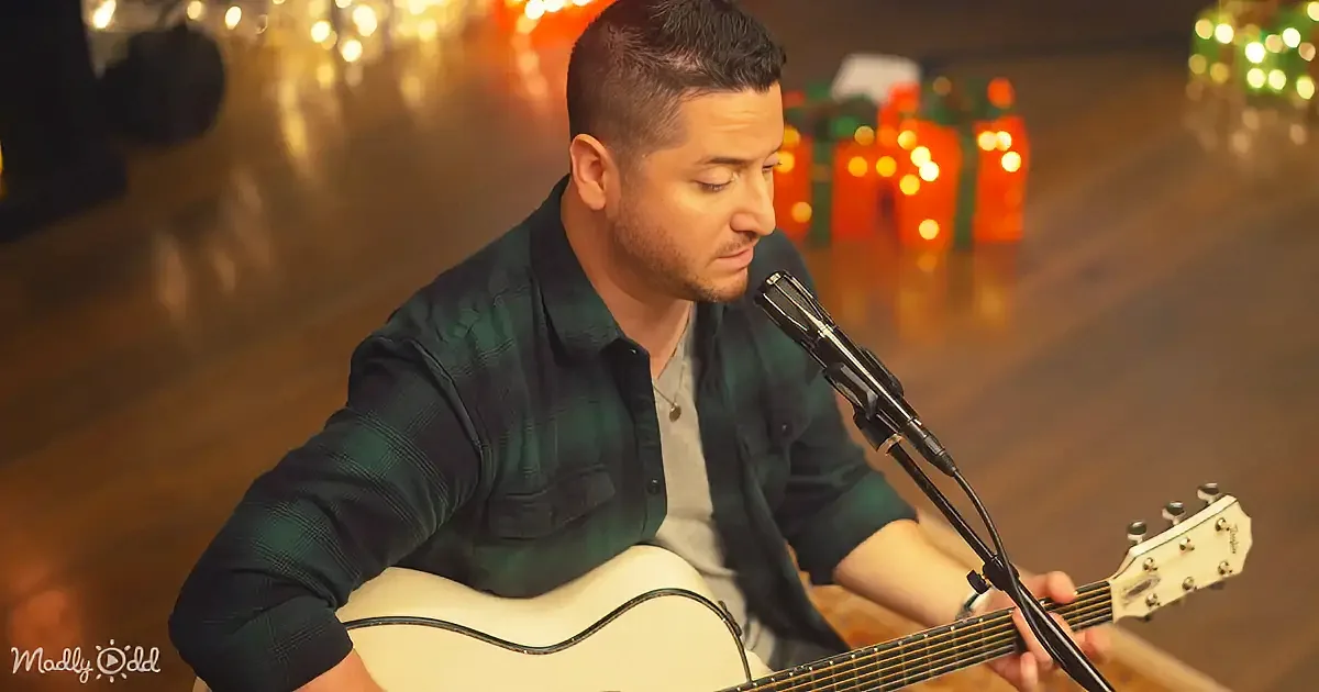 A man in a green plaid shirt plays an acoustic guitar and sings into a microphone.