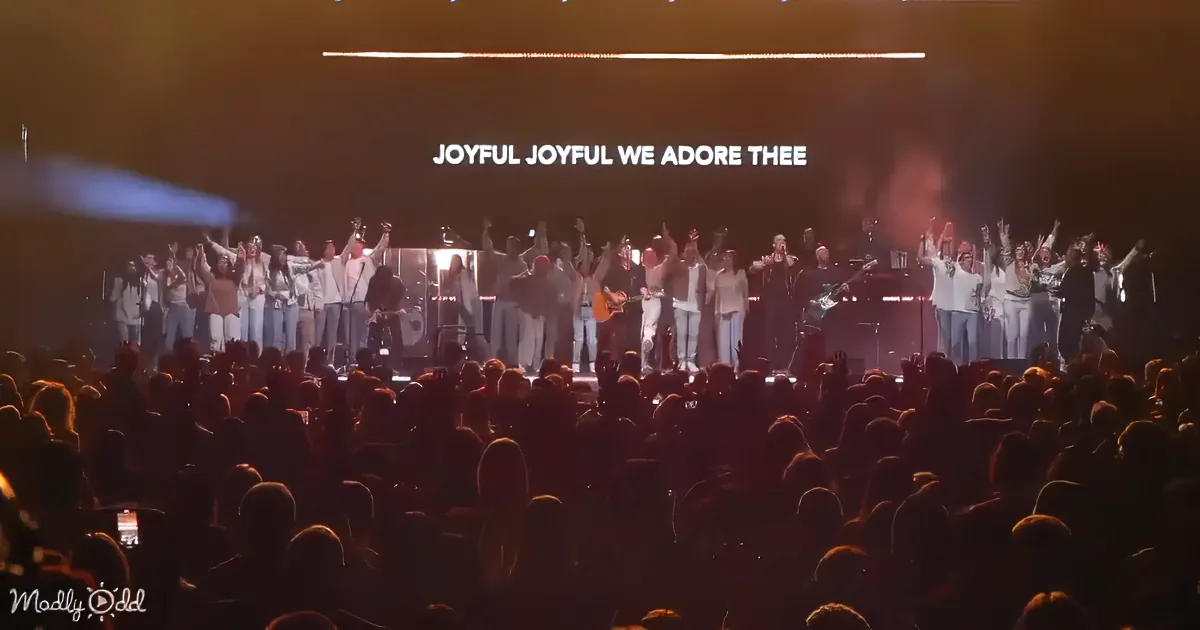 A group of people on stage with arms raised, singing "Joyful, Joyful We Adore Thee," with a large audience in front.