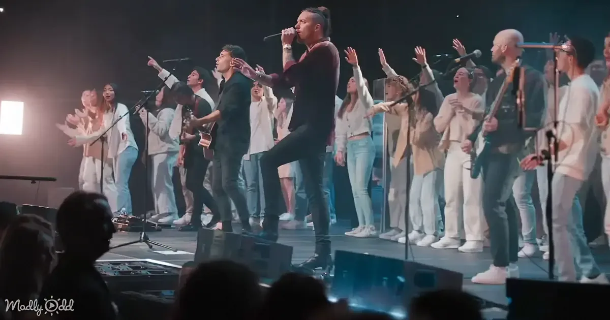 A man in black leads a group of people in worship, raising hands and singing on stage.