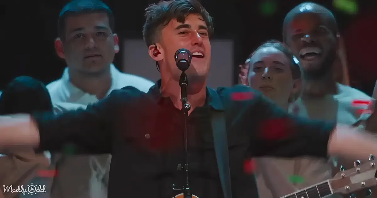 A man in a black shirt sings into a microphone with a group of people behind him.