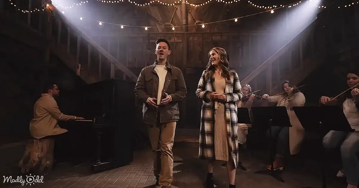 A man and woman sing on stage, accompanied by a pianist and string quartet in a rustic setting.