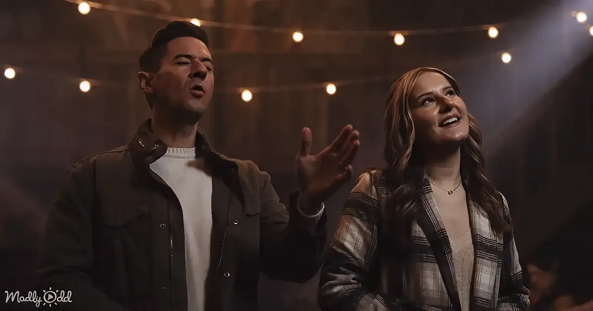 A man and woman are singing passionately, the man gesturing with his hand, both illuminated by warm stage lights.
