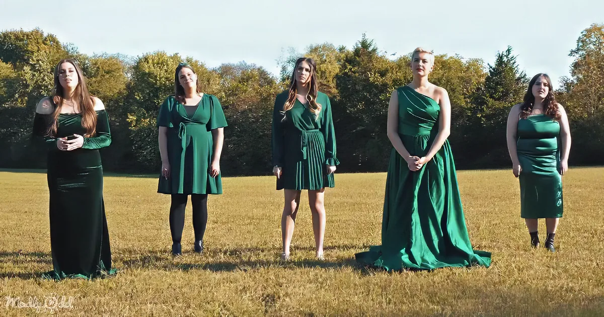 Five women in emerald green dresses stand in a grassy field, posing for a photo.