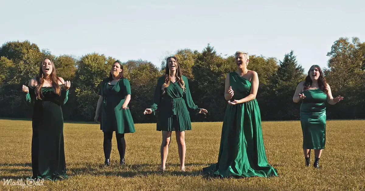 Five women in green dresses sing joyfully in a grassy field with trees in the background.