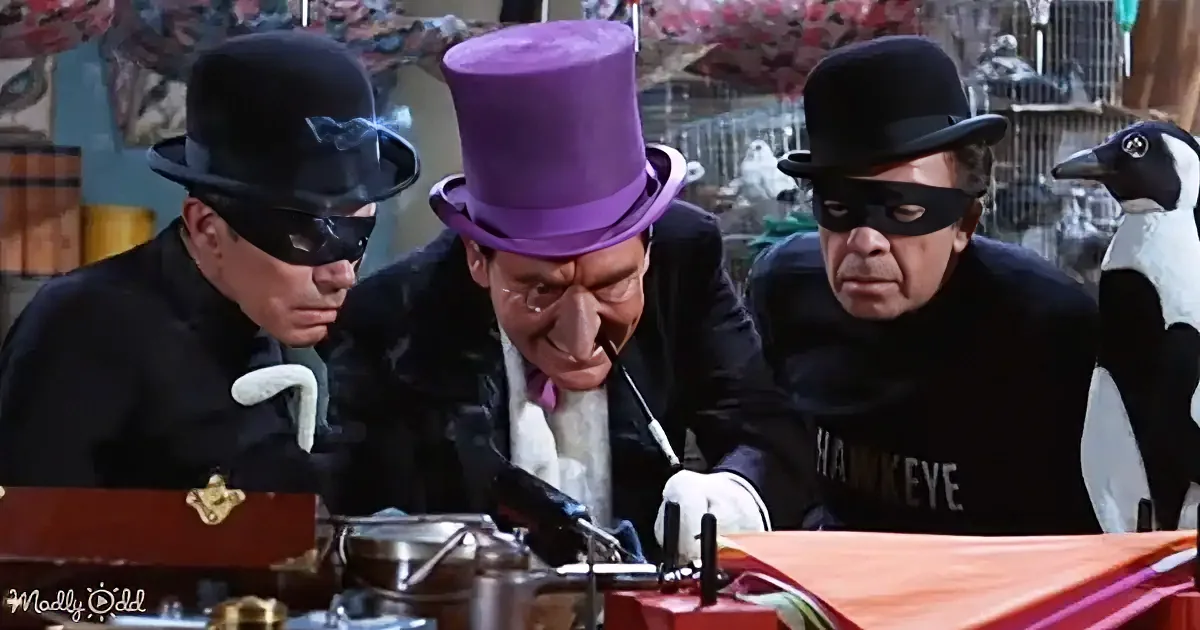 A man in a purple top hat and bow tie is surrounded by two masked figures, examining a device on a table.