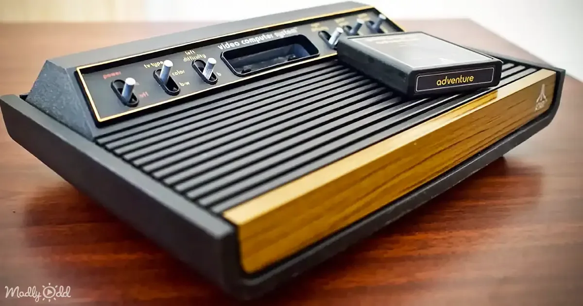 An Atari 2600 video game console with a game cartridge on top is displayed on a wooden surface.