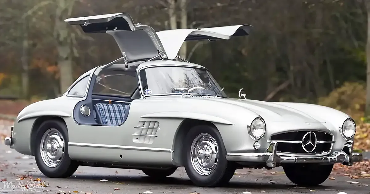 A classic Mercedes-Benz sports car with gull-wing doors open, parked outdoors on a leaf-strewn surface.