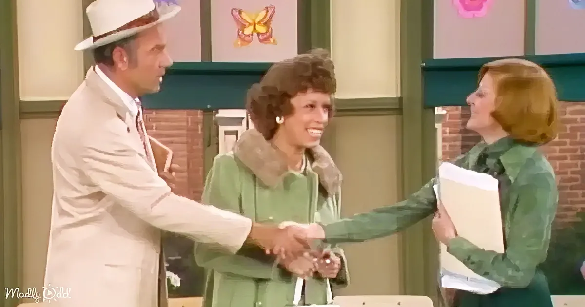 A man in a white suit shakes hands with a woman in a green coat, both smiling, in a colorful room.