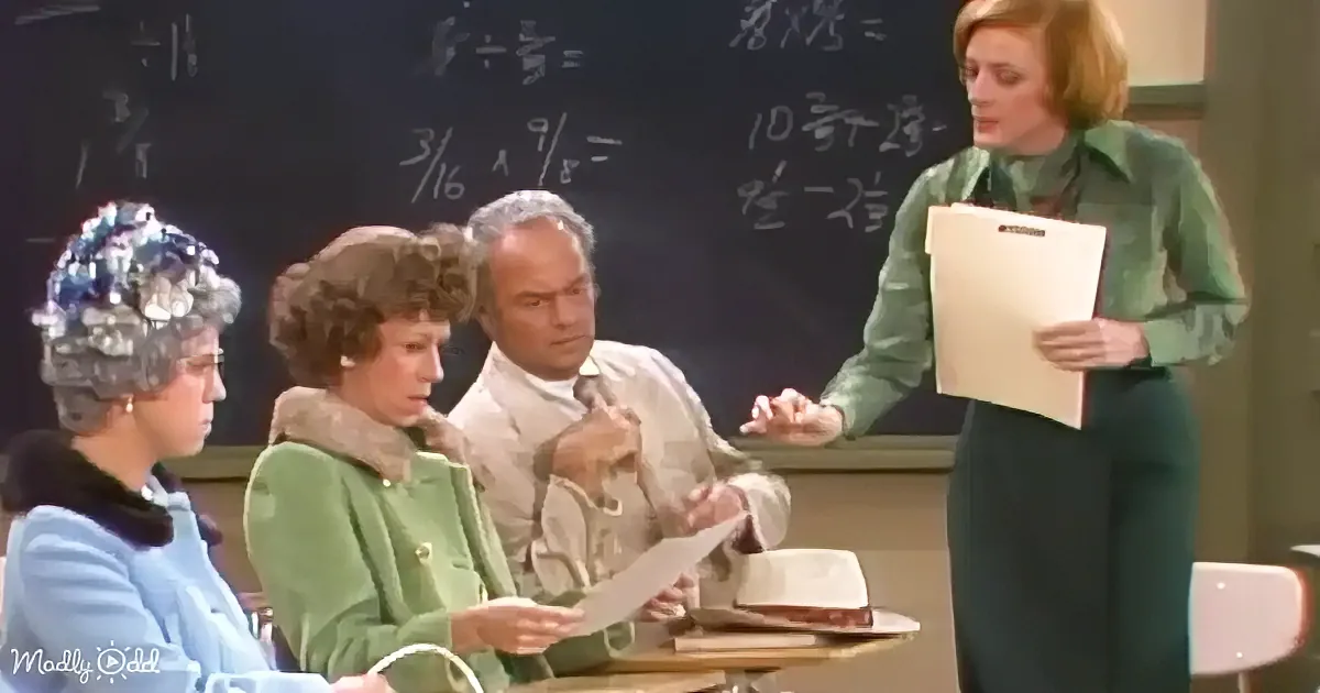 A woman in a green dress holds a clipboard, speaking to a seated audience in front of a chalkboard.