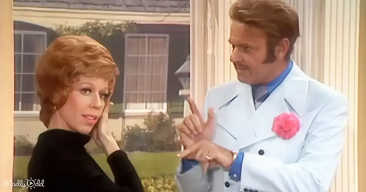 A woman in black adjusts her hair while a man in a white suit with a pink flower gestures beside her.