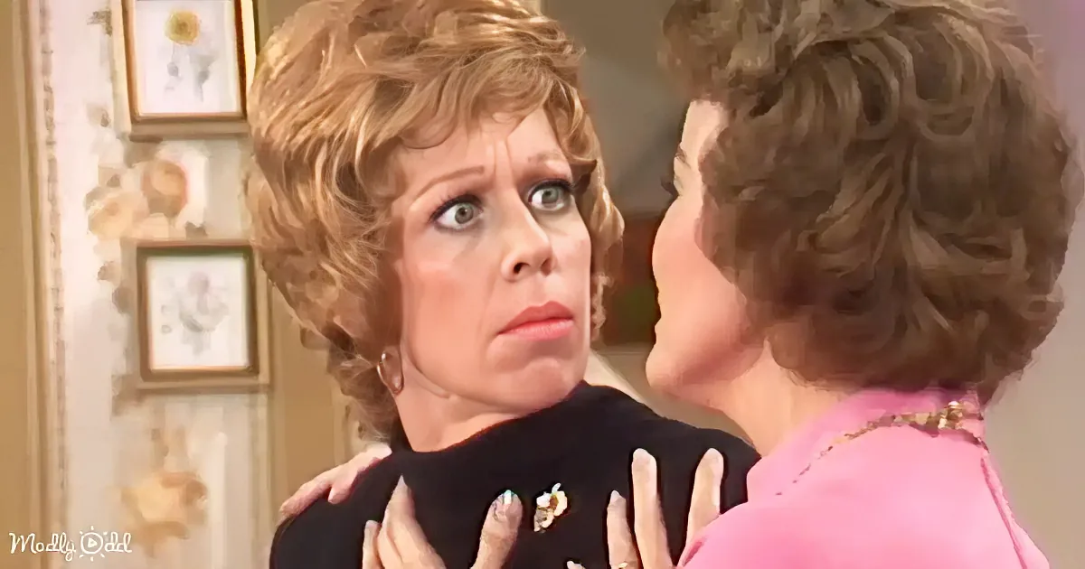 A woman in a black top is being embraced by another woman in a pink jacket in a warmly lit room with framed pictures.