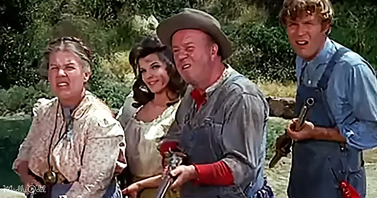 A man in a cowboy hat and overalls holds a guitar, surrounded by three other individuals in a rustic setting.