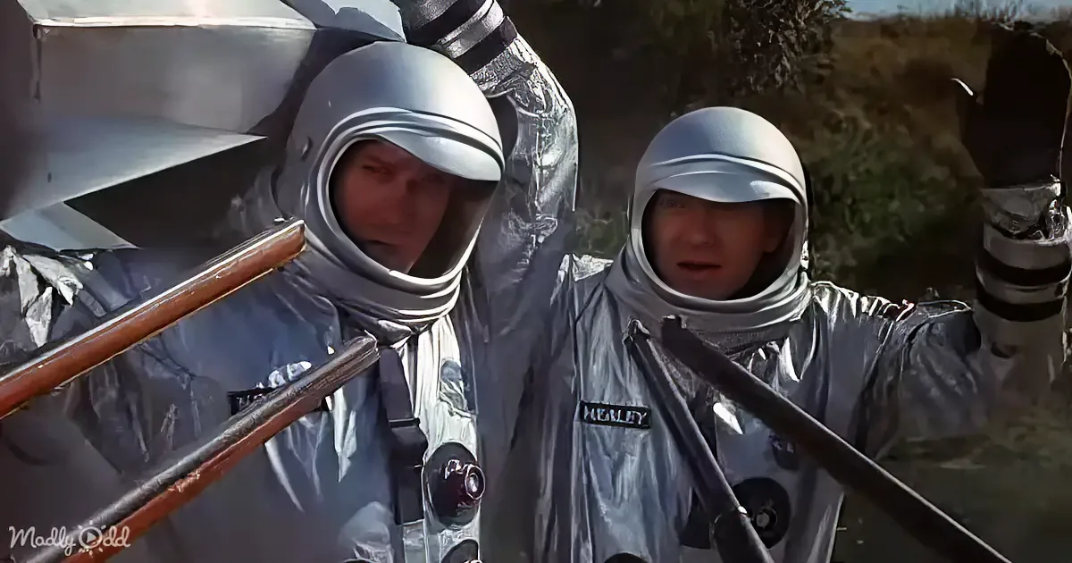 Two astronauts in silver suits, one holding a tool, appear to be working on a spacecraft.