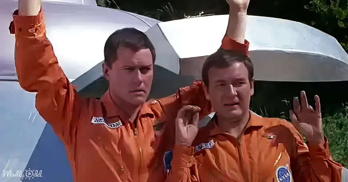 Two men in orange jumpsuits hold up objects, one with a surprised expression, against a white structure and greenery.