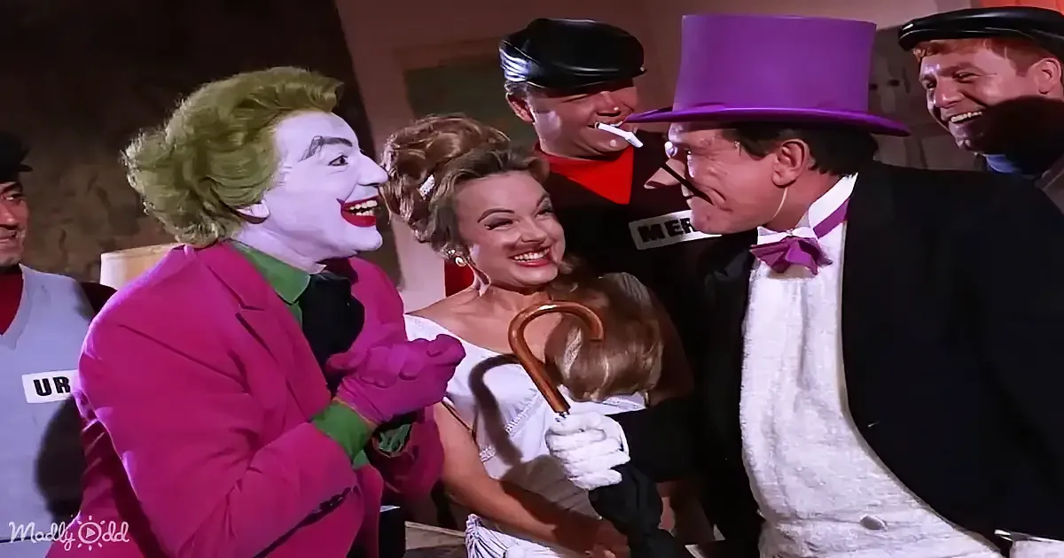 The Joker in a pink suit and green hair is laughing with a woman in a white dress and a man in a purple top hat.