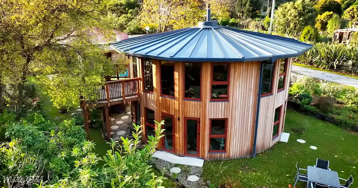 A circular wooden house with a blue metal roof sits surrounded by lush greenery and a stone pathway.