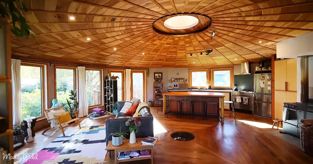 A cozy, sunlit living space with wooden ceiling, floor, and furniture, featuring a kitchen island and large windows.