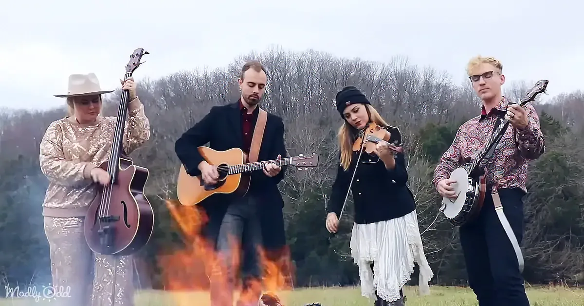 A band plays outdoors near a fire, with a woman in a cowboy hat and a man in a suit among the musicians.