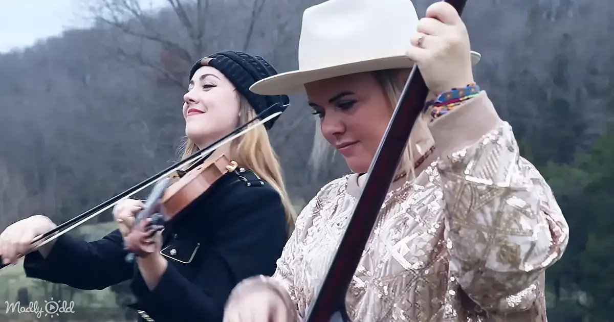 A woman in a sequined shirt plays a cello, another in a black jacket plays a violin, both outdoors.